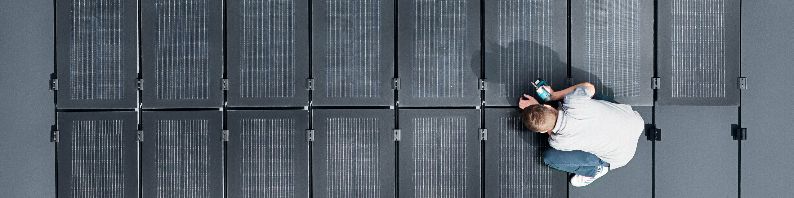 Zwei Personen installieren PREFALZ-Solarmodule auf einer großen grauen Dachfläche, die mit doppelflanschigen Paneelen bedeckt ist.