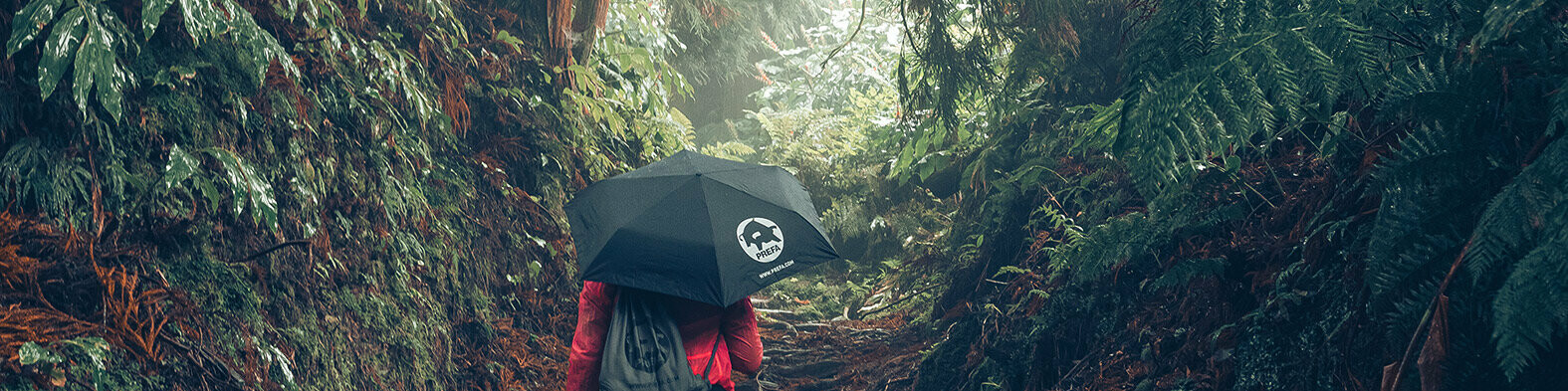 Image of a forest with a female hiker in a red jacket with a PREFA umbrella and gym bag, symbolises PREFA’s environmental protection and sustainability, as well as the closed-loop economy and recycling